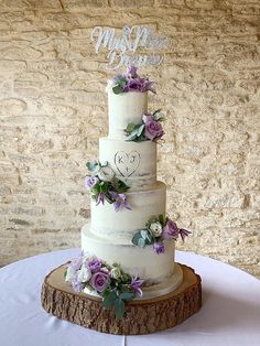 a three tiered cake with purple flowers and greenery on the top is displayed in front of a stone wall