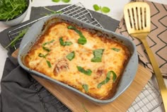 a casserole dish with cheese and spinach on a wooden cutting board next to a fork