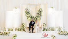 a bride and groom sitting on a chair in front of a floral decorated backdrop at their wedding