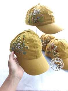 three hats with embroidered flowers on them, one being held up to the side by a person's hand