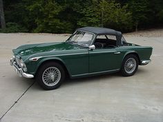 an old green sports car parked in a parking lot with trees in the back ground