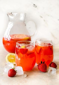 two glasses filled with red liquid and strawberries next to lemon wedges on a marble surface