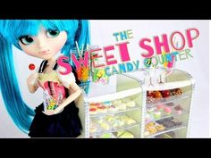 a doll with blue hair holding a cupcake in front of the sweet shop candy counter