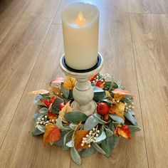 a white candle is sitting on top of a wreath with leaves and berries around it