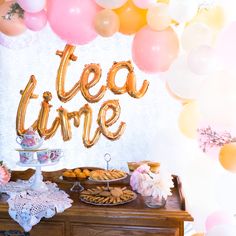 there is a tea time sign on the table with desserts and balloons in the background