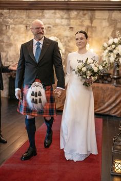 a man and woman walking down a red carpeted aisle holding hands while dressed in kilts