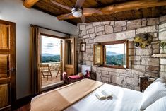 a bedroom with a stone wall and ceiling fan in front of a window looking out onto the mountains