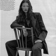 a woman sitting on top of a wooden chair in front of a white wall and wearing a suit