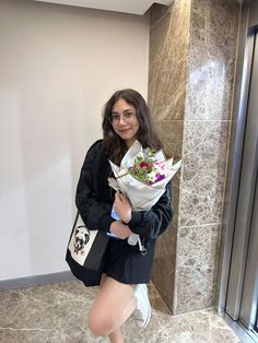 a young woman is holding flowers in her hand and posing for the camera while standing next to a wall