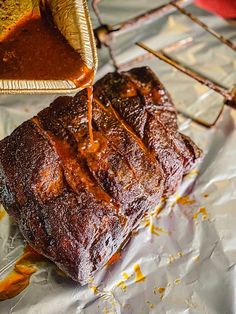 a person pouring sauce onto a piece of meat on tin foil with other food items in the background