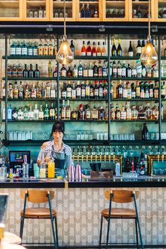 a woman sitting at a bar with lots of bottles on the wall behind her and two chairs in front of it