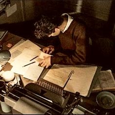 a man sitting at a desk writing on a piece of paper next to an old typewriter