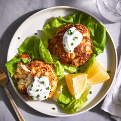 two crab cakes on lettuce with sour cream and lemon wedges next to silverware