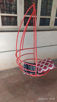 a red metal swing chair with a cushion on the ground in front of a window