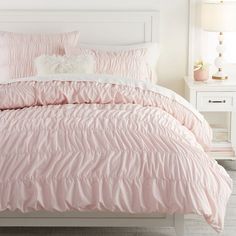 a bed with pink comforter and pillows in a white room next to a night stand