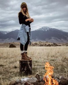 a woman standing on top of a tree stump next to a campfire with mountains in the background