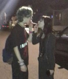 two people standing next to each other in front of a car on a street at night