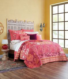 a bed with pink and orange comforters in a bedroom next to a window on a hard wood floor