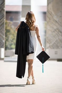 a woman walking down the street with her graduation cap and gown in one hand, she is holding a black jacket over her shoulder