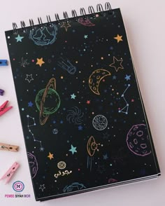 an open notebook with space drawings on it and colored pencils next to the pages