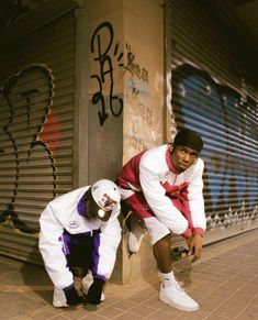 two young men leaning against a wall with graffiti on the walls behind them, both wearing white and purple outfits