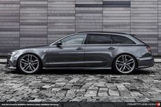 a grey car parked in front of a building on a cobblestone street with a brick wall behind it