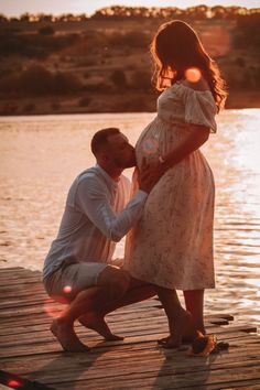 a man kneeling down next to a woman on a dock near the water at sunset