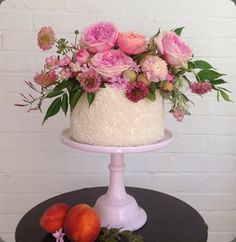 a white cake with pink flowers and peaches on a table next to a brick wall