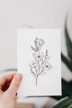 a person holding up a card with flowers on it in front of a potted plant