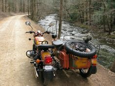 an orange motorcycle parked on the side of a dirt road next to a river and trees