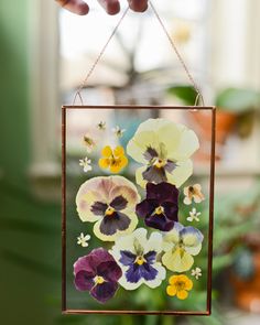 a person holding up a small glass frame with flowers on it in front of a potted plant