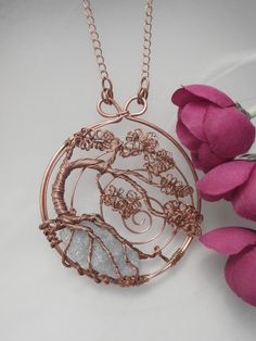 a necklace with a tree on it and pink flowers in the foreground, sitting next to a white surface