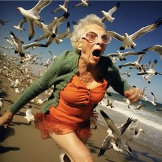 an old woman running on the beach with seagulls flying around her