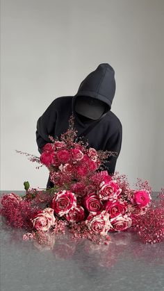 a person in a black hoodie is surrounded by pink flowers on a gray surface