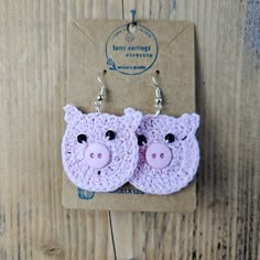 two pink crocheted pig earrings hanging from hooks on a wooden background with a card