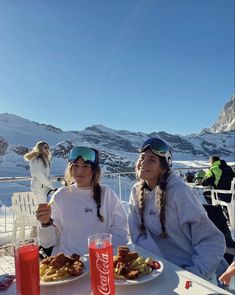 two women sitting at a table with food and drinks in front of snow covered mountains