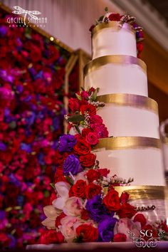 a wedding cake with red and purple flowers on the bottom tier is decorated with gold ribbon