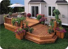 a wooden deck with potted plants and flowers on the bottom level, in front of a house