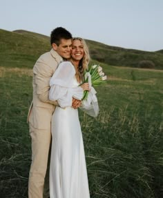 a bride and groom standing in a field with their arms around each other holding flowers