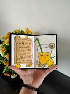 a hand holding up an open book with a drink inside it and some flowers in the background