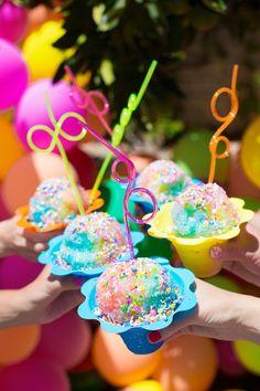 hands holding small cupcakes with sprinkles on them in front of balloons