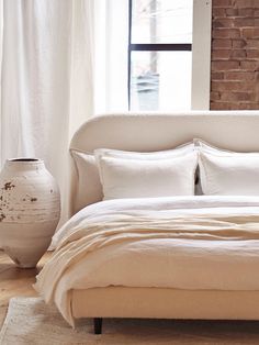 a bed with white linens and pillows in front of a brick wall next to a window