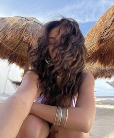 a woman sitting on top of a sandy beach next to straw umbrellas with her arms crossed