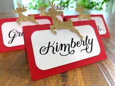 three red and white christmas place cards with gold reindeer on them, sitting on a wooden table