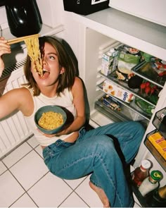 a woman sitting in front of an open refrigerator holding a bowl of macaroni and cheese