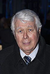 an older man with white hair wearing a black coat and tie, looking at the camera