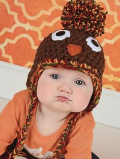 a baby wearing a knitted owl hat with big eyes and an orange shirt is sitting on the floor