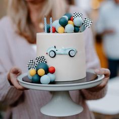 a woman is holding a cake decorated with cars and balloons on it while wearing a white sweater