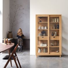 a woman sitting at a table in front of a wooden cabinet filled with dishes and cups