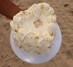 a white cake with shells and gold decorations on it sitting in the sand near a person's hand